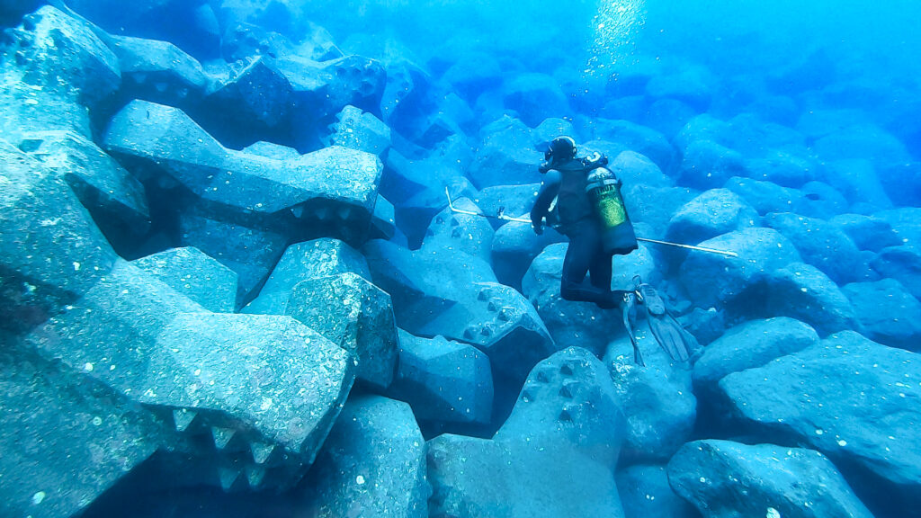 Expert CLAS inspectant la sous-couche d’une digue maritime à travers la carapace en blocs artificiels à l’aide d’un outil de mesure et de prise de vue sous-marine